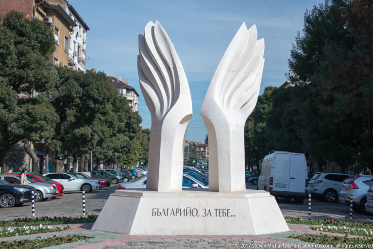 Paired white sculptural forms on an inscribed pedestal, surrounded by parked cars, trees, and apartment buildings in Sofia.