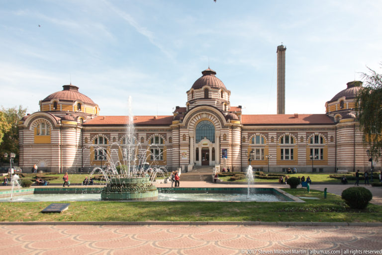 Sofia’s former Central Mineral Baths with striped façade, domed pavilions, central entrance, fountains, and landscaped grounds.