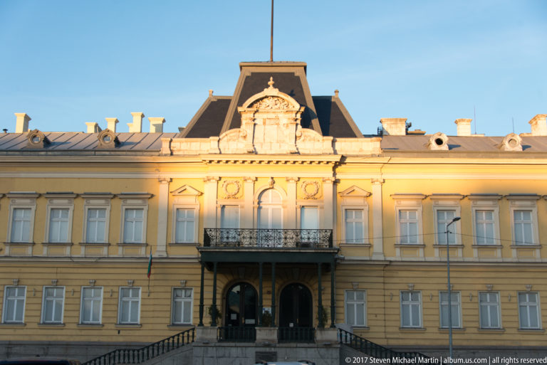 Symmetrical yellow historic building with central balcony, arched entrance, sculpted pediment, slate roof, and Bulgarian flag.