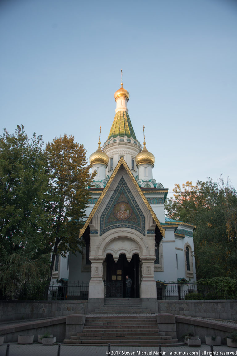 Front view of Sofia’s Russian Church with gabled entrance porch, white walls, green roofs, and golden onion domes.