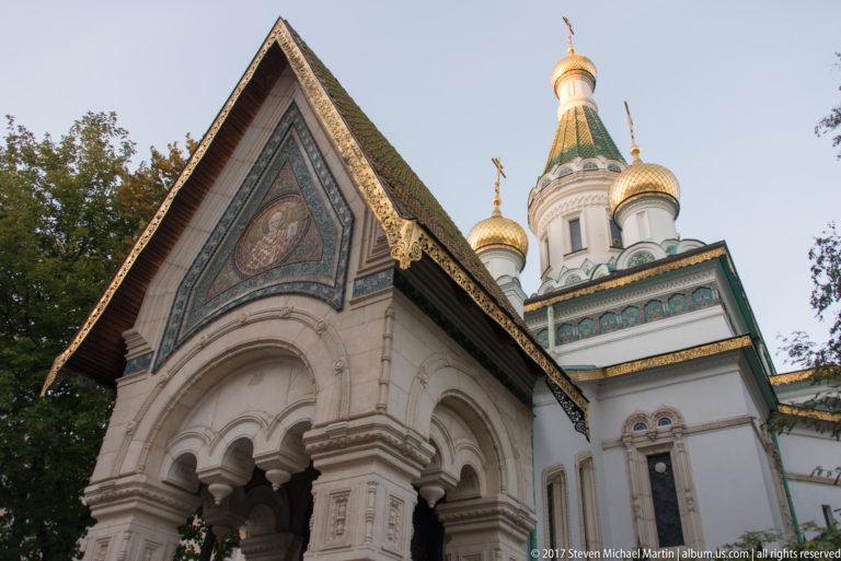 Angled view of the Russian Church entrance porch with carved arches, steep gable, white drums, and golden onion domes.