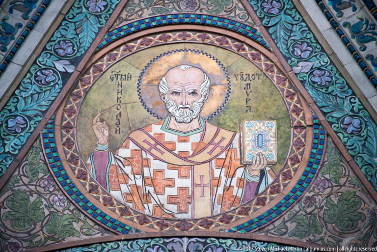 Round mosaic of Saint Nicholas as a haloed bishop holding a Gospel book, surrounded by ornate turquoise and gold patterns.