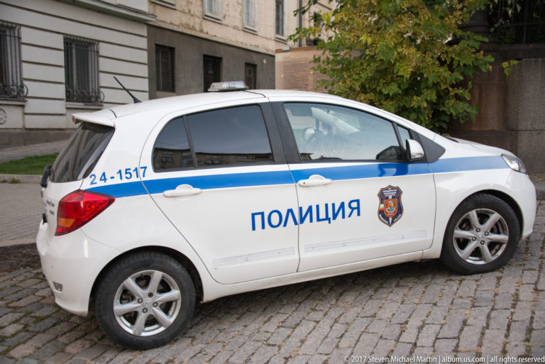White Bulgarian police hatchback with blue stripe, crest, unit number, and Cyrillic police lettering parked on cobblestones.