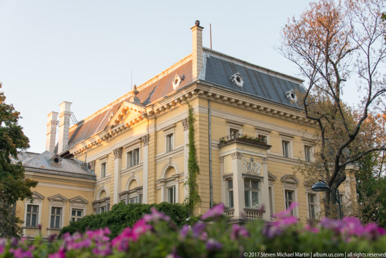 Historic pale-yellow mansion with ornate window surrounds, gray dormered roof, ivy, and purple flowers in the foreground.