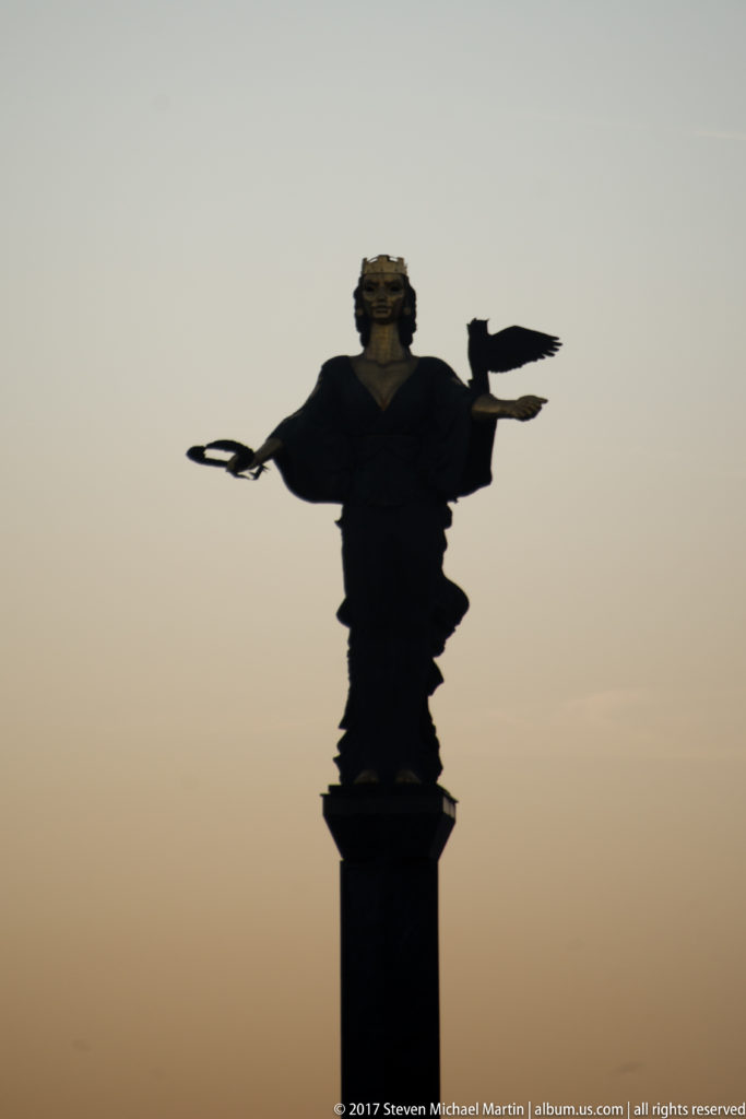 Silhouetted Statue of Saint Sofia high on its column, with outstretched arms and symbolic attributes against the evening sky.