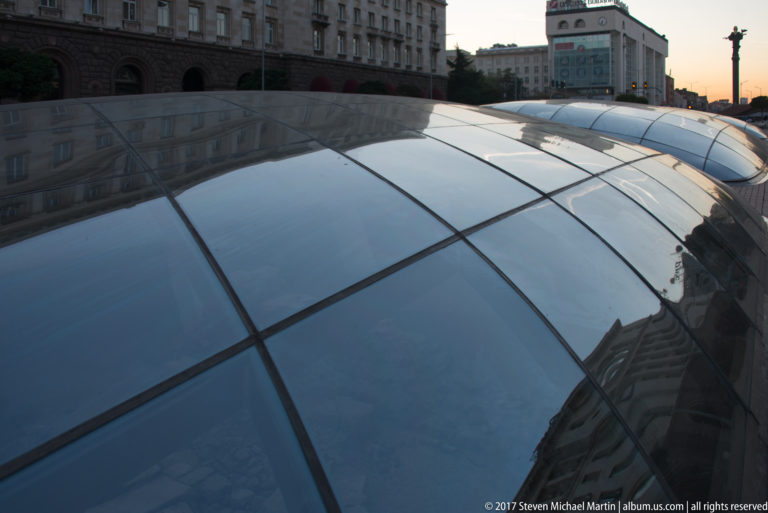 Curved glass canopy over Sofia’s central Serdica archaeological area, reflecting surrounding buildings in the evening light.