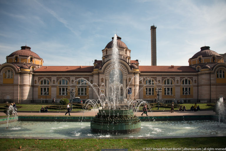 Fountain jets in front of Sofia’s former Central Mineral Baths and its domed, striped historic façade.