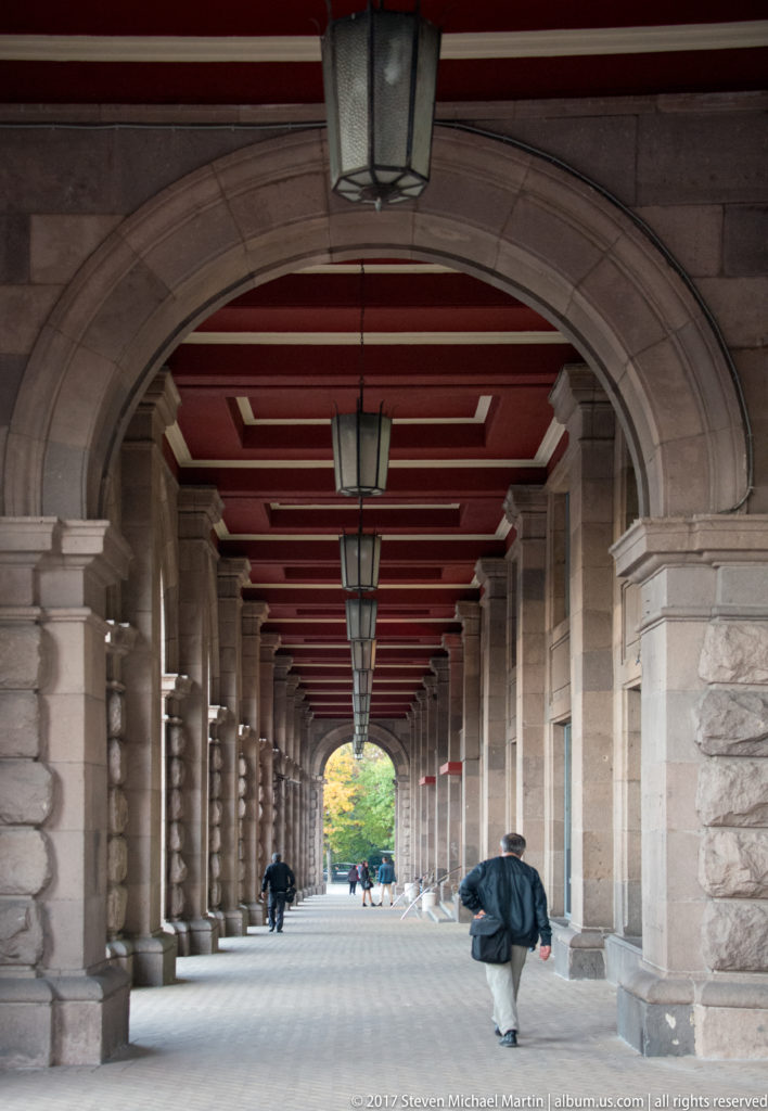 Long stone arcade with repeated arches, red ceiling beams, hanging lanterns, and pedestrians receding toward daylight.