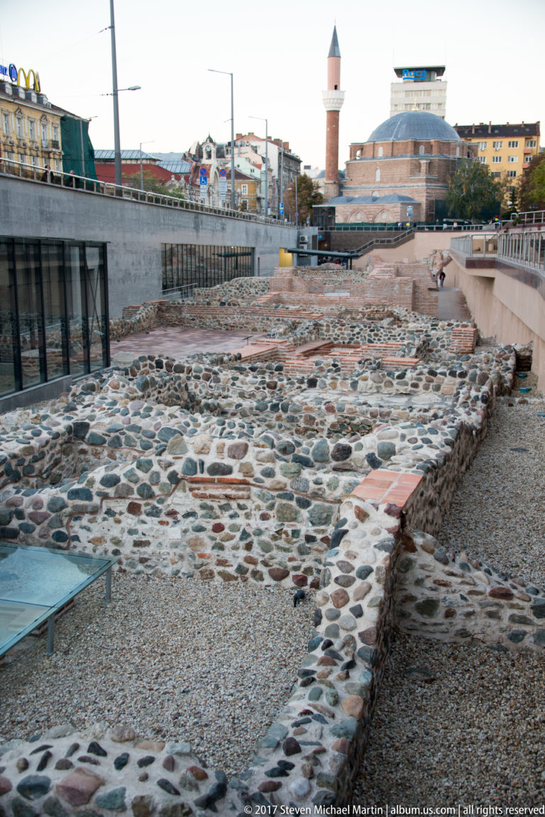 Excavated stone foundations of ancient Serdica with gravel paths, modern buildings, and a mosque minaret in the background.