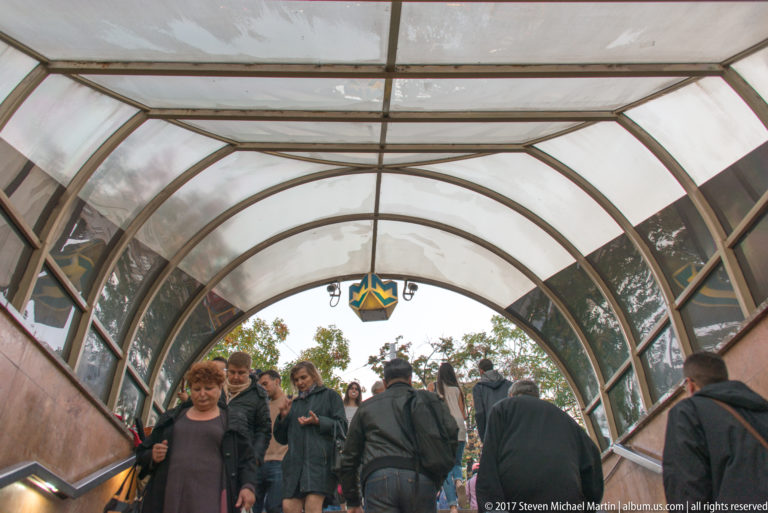 Crowd of pedestrians moving through a Sofia Metro entrance beneath a curved transparent canopy.