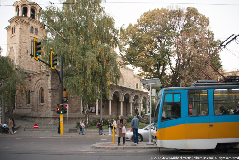 Blue-and-yellow Sofia tram crossing an intersection with pedestrians, traffic lights, overhead wires, and historic stone arcades.