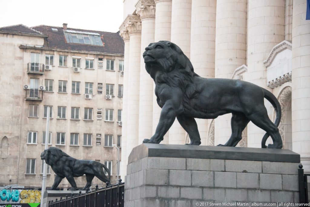 Large bronze lion on a stone pedestal beside the columned Palace of Justice, with a second lion visible behind.