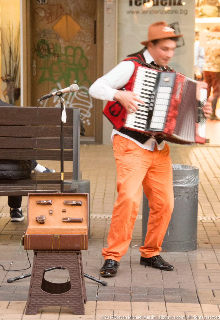 Street musician playing a red-and-white accordion beside an open tip suitcase and microphone stand on a pedestrian street.
