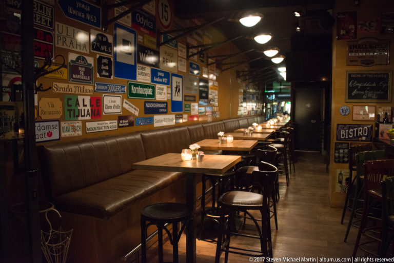 Long narrow bistro interior with banquette seating, small tables, stools, candles, and walls densely covered in vintage signs.