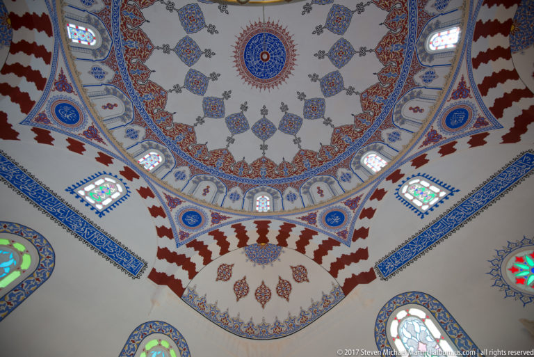 Ornately painted dome and pendentives with blue, red, and gold motifs and small stained-glass windows inside a Sofia mosque.