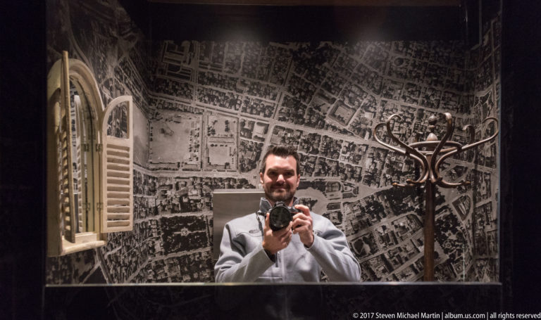 Man holding a camera reflected in a mirror before a black-and-white city map mural and coat rack.