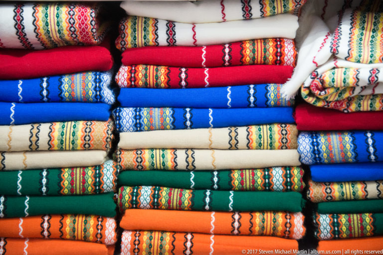 Stacks of folded red, blue, green, orange, and cream textiles decorated with repeated multicolored geometric folk patterns.