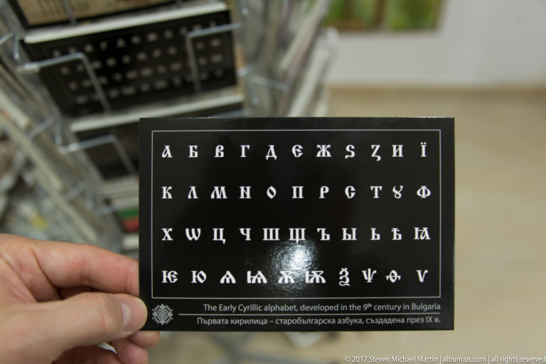 Hand holding a black card printed with early Cyrillic characters and explanatory English and Bulgarian text.