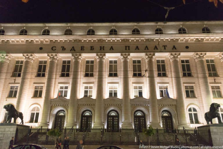 Illuminated Palace of Justice in Sofia with monumental columns, arched entrances, Bulgarian inscription, and lion sculptures at night.