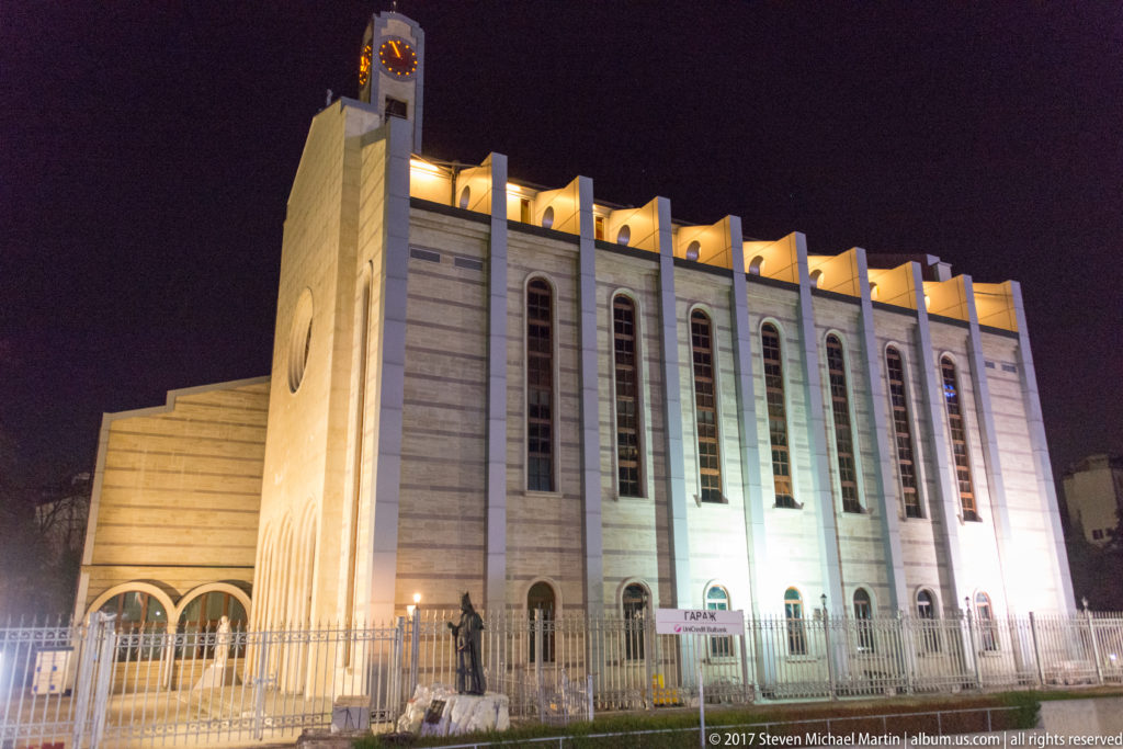 Night view of the modern Cathedral of St. Joseph with clock tower, tall arched windows, circular opening, and illuminated roofline.
