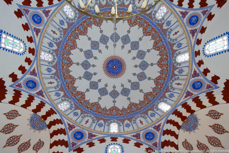Centered view of a mosque dome with concentric blue, red, and gold ornament and stained-glass windows around its base.