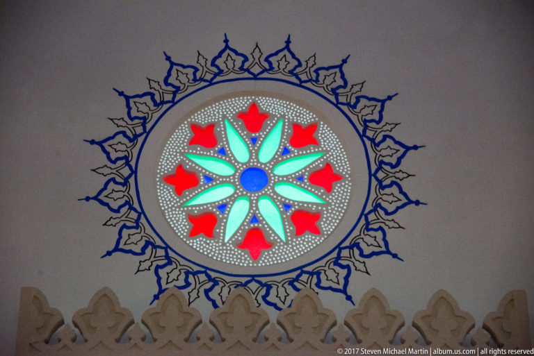 Circular stained-glass rosette with red, turquoise, and blue floral geometry on a pale mosque wall.
