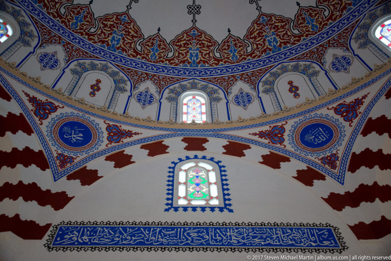 Painted mosque arches with blue Arabic calligraphy medallions, floral ornament, and small stained-glass windows.