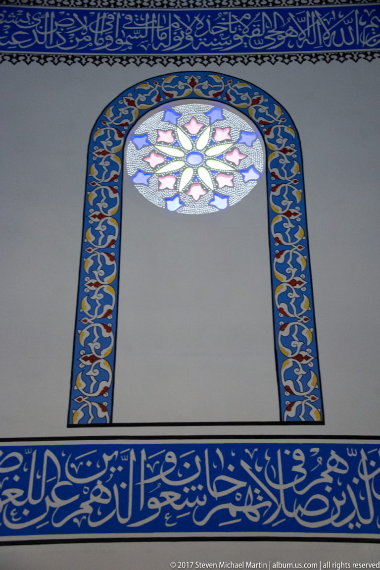 Small arched stained-glass window surrounded by blue-and-gold ornament and horizontal Arabic calligraphy bands.