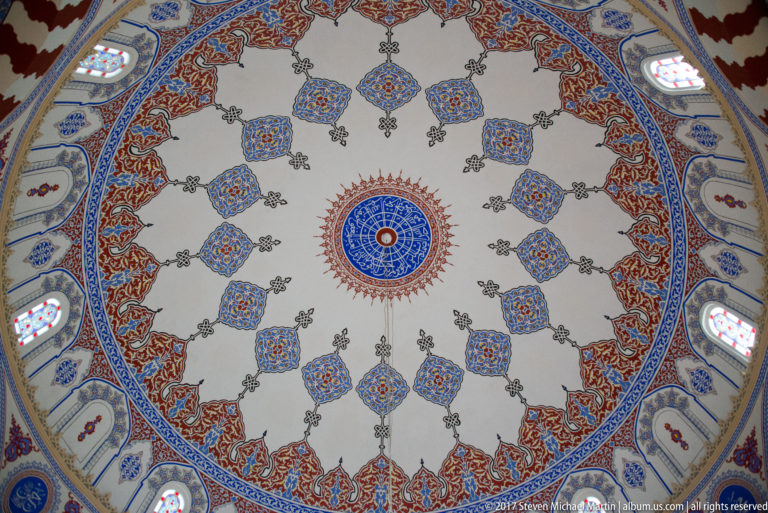 Upward close view of a mosque dome covered with repeating blue, red, green, and gold pendant-like floral motifs.
