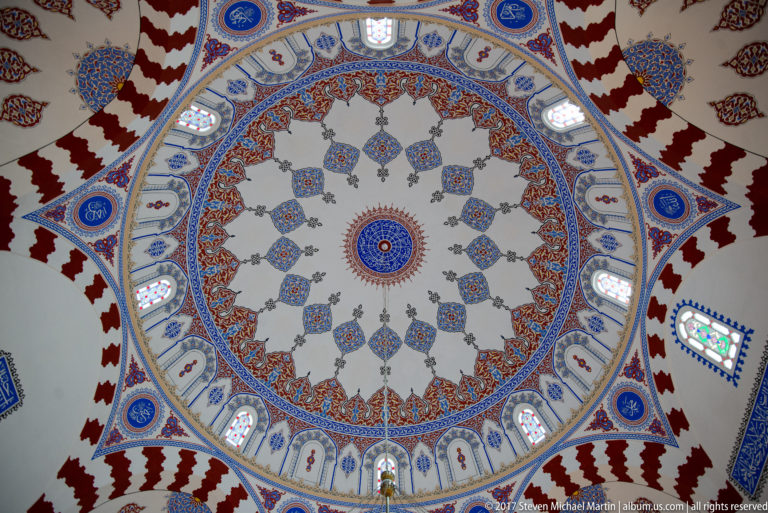 Oblique upward view of a painted mosque dome, pendentives, red-and-white bands, blue medallions, and stained-glass windows.