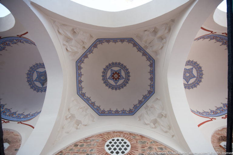 White arches framing decorated ceiling bays with blue-and-red rosettes, sculpted details, exposed brick, and a round grille.
