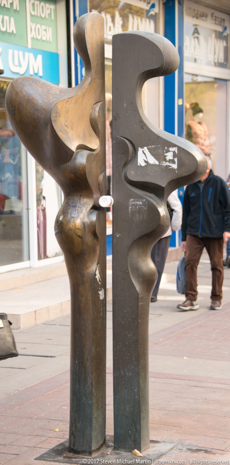 Abstract bronze-toned and dark metal sculpture with interlocking curved forms on a pedestrian street in Sofia.