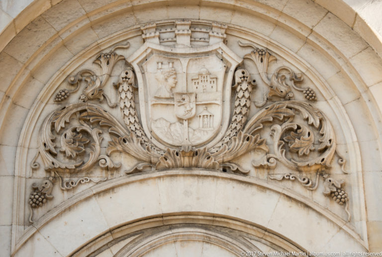 Carved stone coat of arms above an arched entrance, surrounded by scrollwork, foliage, and grape-cluster motifs.