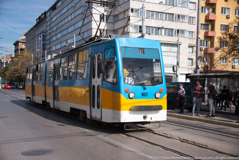 Yellow, white, and blue Sofia tram traveling on street tracks beneath overhead wires with pedestrians and apartment buildings nearby.