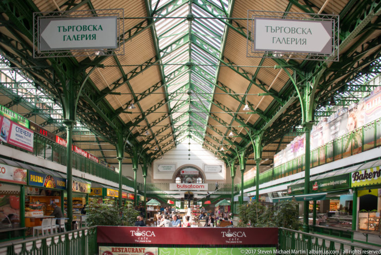 Interior of Sofia’s Central Market Hall with green iron trusses, glazed roof, shop signs, food counters, and restaurant seating.