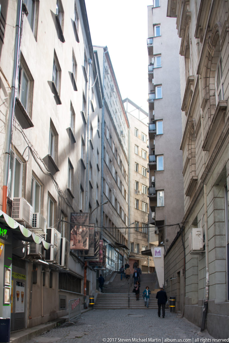 Narrow Sofia side street between mid-rise buildings with air conditioners, service entrances, blank walls, and pedestrians in the distance.