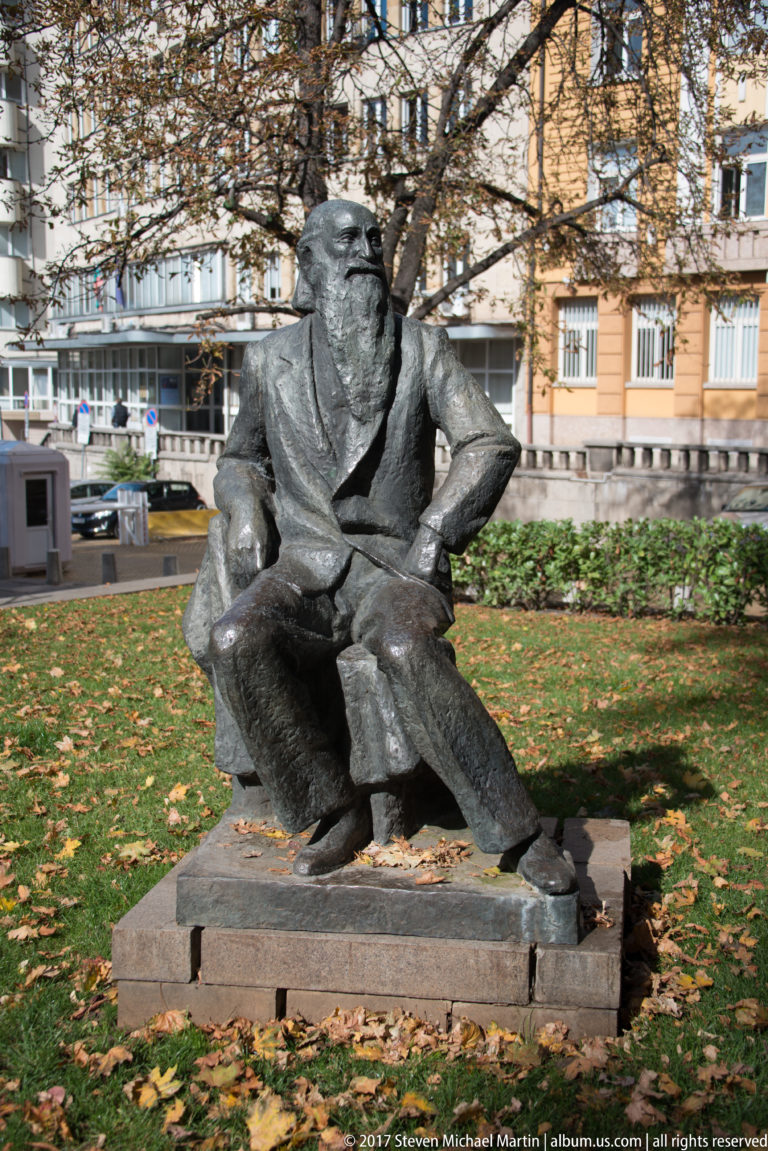 Dark bronze sculpture of a seated figure in a Sofia park with autumn leaves, trees, lawn, and city buildings behind.