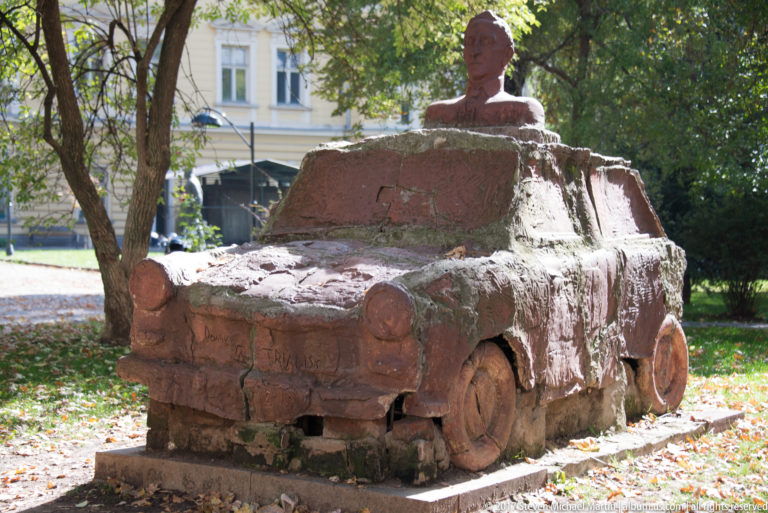 Reddish park sculpture shaped like a car with a portrait bust emerging from the roof, surrounded by trees and grass.