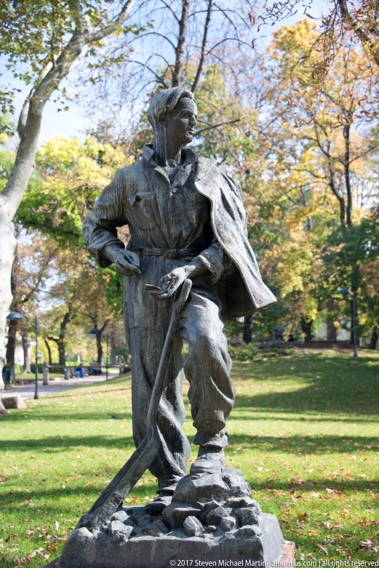 Dark sculpture of a cloaked figure crouching with a long sword or staff in a tree-filled Sofia park.