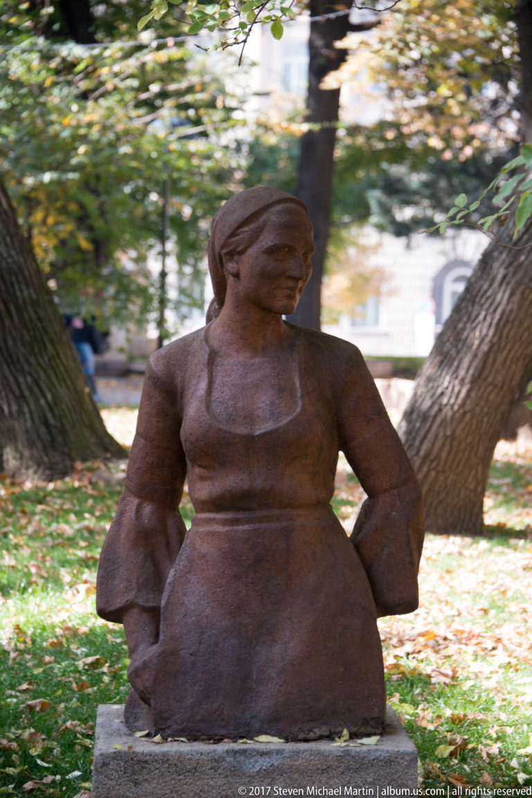 Reddish-brown sculpture of a seated woman with simplified forms beneath mature trees in a Sofia park.