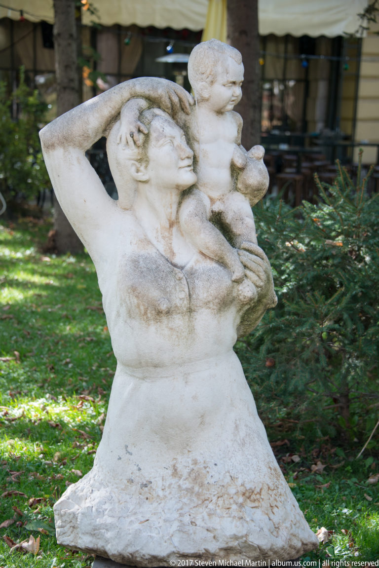 Weathered white sculpture of intertwined human figures with rounded forms in a leafy Sofia park.