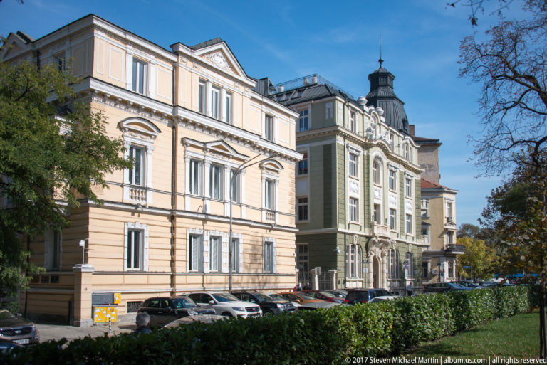 Cream and pale yellow historic buildings with ornate windows, cornices, balconies, and a small tower-like roof feature on a Sofia street.