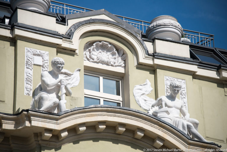 Two white allegorical sculptures seated above an arched window on an ornate pale-green historic façade in Sofia.