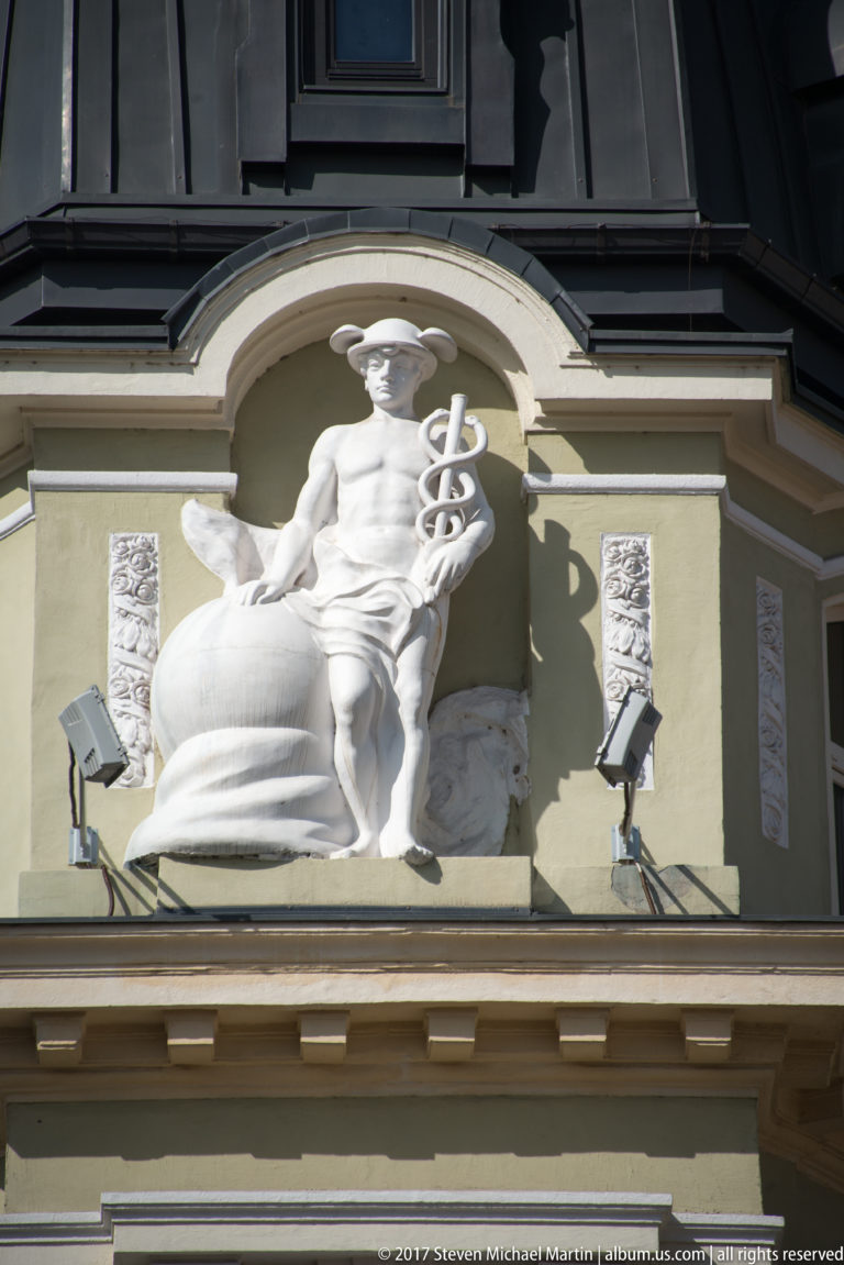 Seated classical figure with a caduceus-like staff mounted high on an ornate historic building façade.