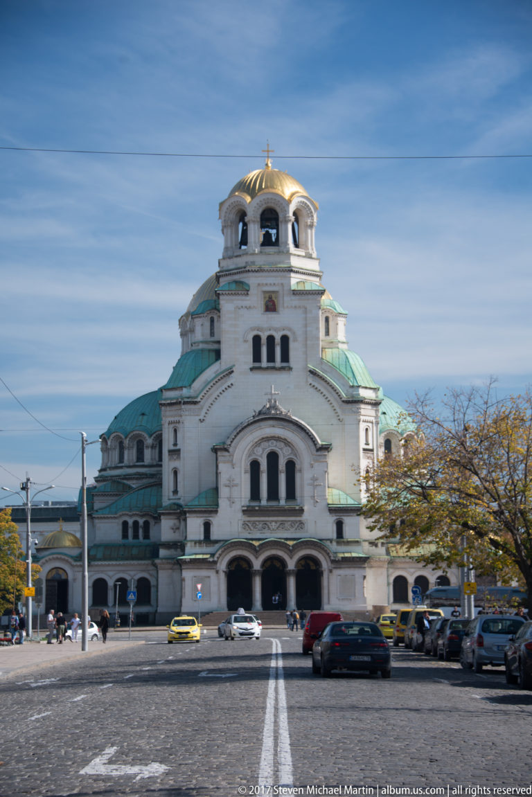 Alexander Nevsky Cathedral viewed frontally from the street, with white walls, green roofs, and a gold-topped tower.