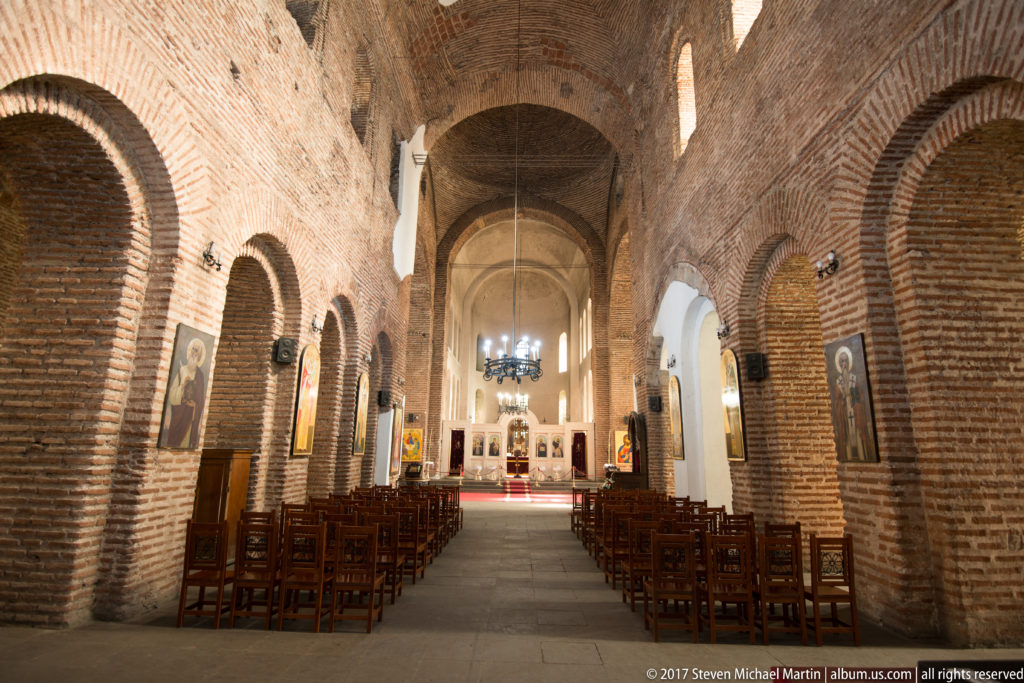 Brick nave of the Church of Saint Sofia with wooden chairs, round arches, icons, and a distant sanctuary.