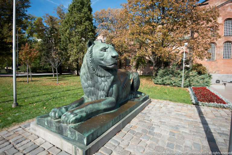 Large reclining bronze lion on a stone pedestal beside a landscaped path with autumn trees and brick architecture.