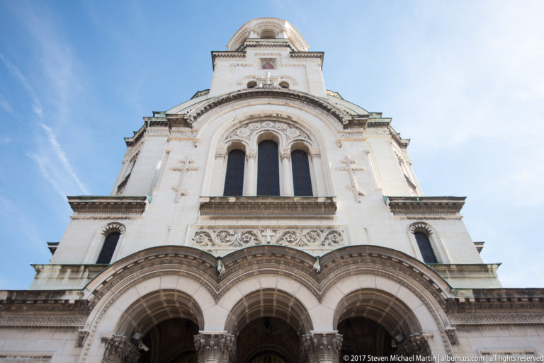 Low-angle view up the main façade of Alexander Nevsky Cathedral, showing triple arches, carved stonework, and the tower above.