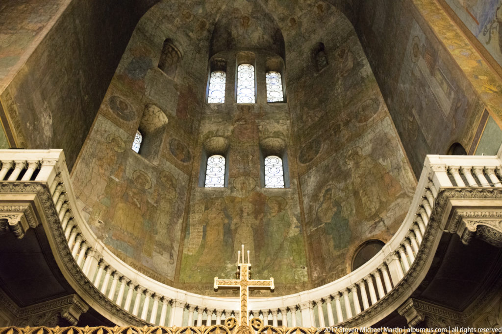 Curved apse interior with religious mosaics, arched windows, pale balustrade, and a cross at Alexander Nevsky Cathedral.