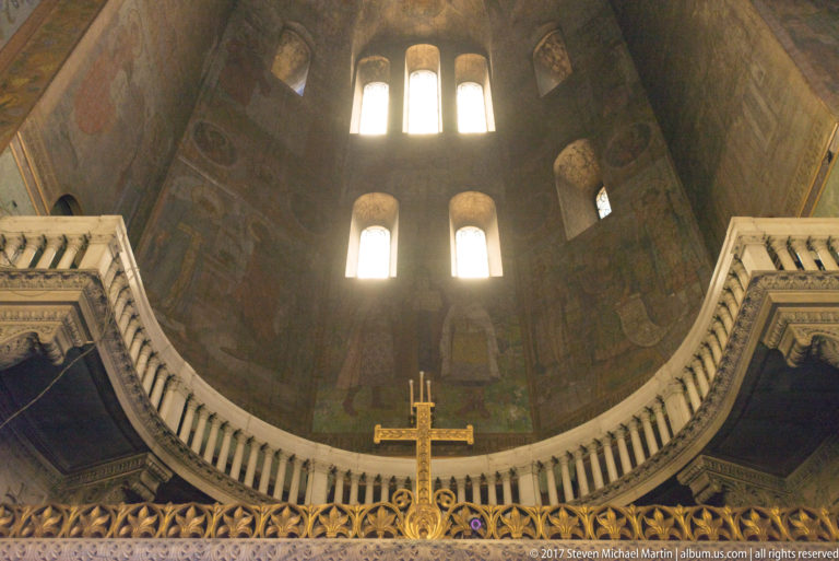 Bright arched apse windows above dark mural-covered walls, a pale balustrade, and gilded cross inside Alexander Nevsky Cathedral.